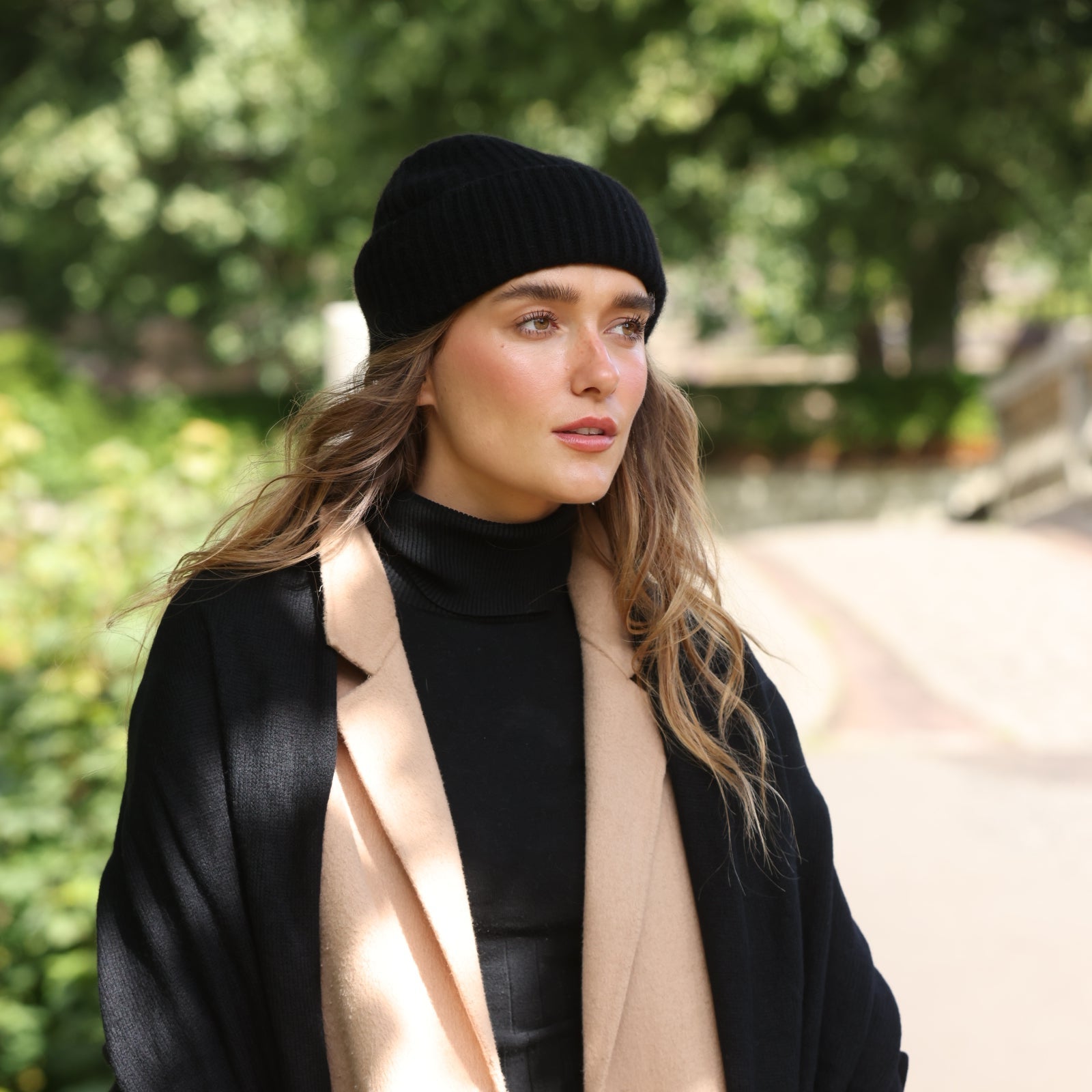 Woman wearing a black beanie and coat outdoors with greenery in the background