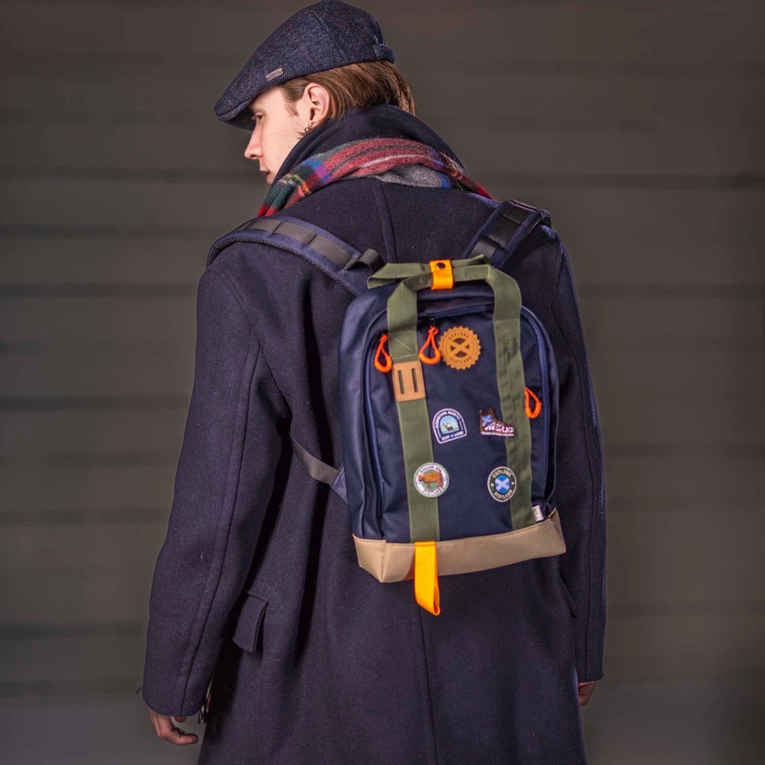 Person wearing a navy coat and cap with a navy blue backpack featuring patches on a grey background