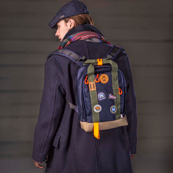 Person wearing a navy coat and cap with a navy blue backpack featuring patches on a grey background
