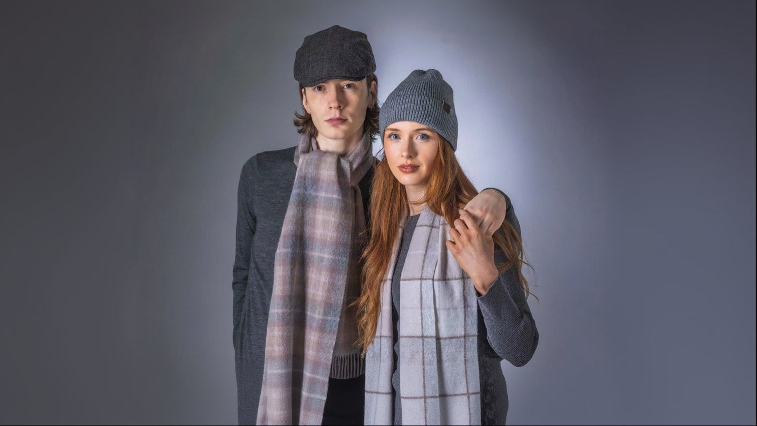Two models posing together wearing knit beanies and plaid scarves against a dark gray studio background.