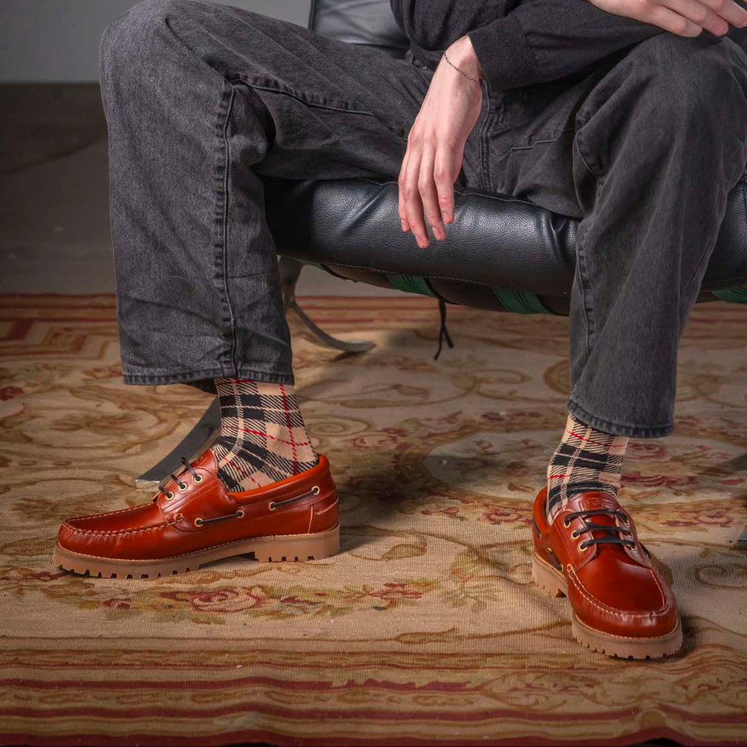 Man sitting on a black leather chair wearing a dark sweater, gray pants, and red shoes.