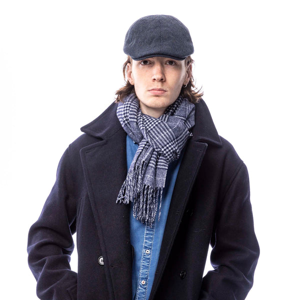 A person wearing a flat cap with a herringbone tweed pattern. They are also wearing a scarf and a coat, suggesting a stylish yet casual look.