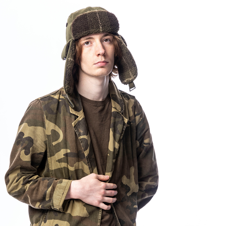 A young man wearing a brown and green camouflage jacket, a dark brown t-shirt, and a matching camouflage and brown faux-fur trapper hat, photographed against a solid white background.