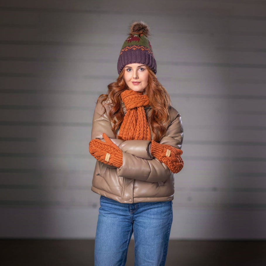 A woman stands with arms crossed wearing a brown quilted jacket, a chunky orange knit scarf and matching knit mittens, a purple knit beanie with a pom-pom, and blue jeans against a gray studio background.