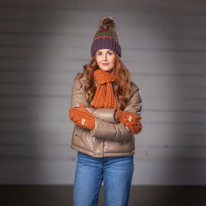A woman stands with arms crossed wearing a brown quilted jacket, a chunky orange knit scarf and matching knit mittens, a purple knit beanie with a pom-pom, and blue jeans against a gray studio background.