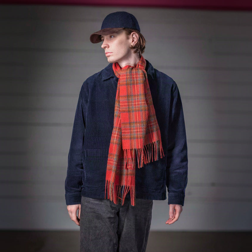 A model wearing a navy blue corduroy jacket, dark gray pants, a navy cap, and a red and green plaid scarf, standing against a gray paneled background.