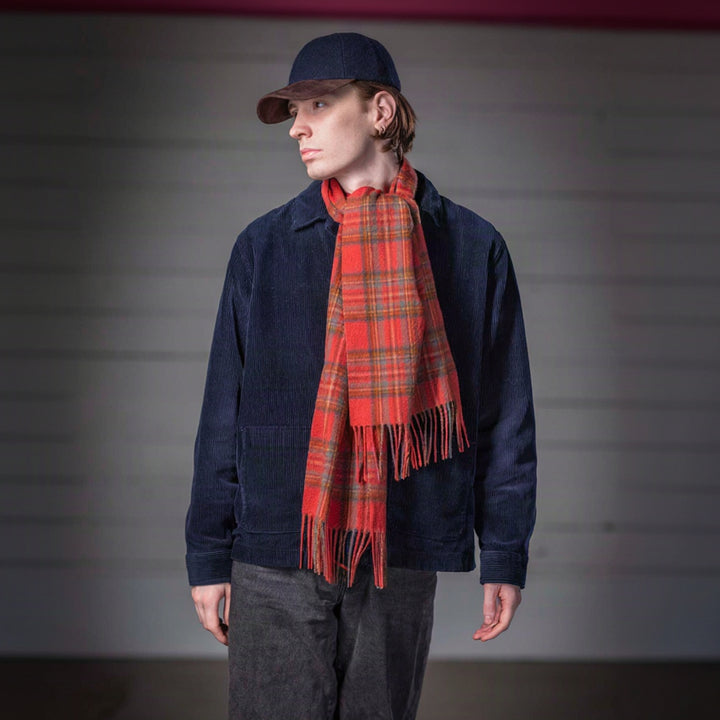 A model wearing a navy blue corduroy jacket, dark gray pants, a navy cap, and a red and green plaid scarf, standing against a gray paneled background.