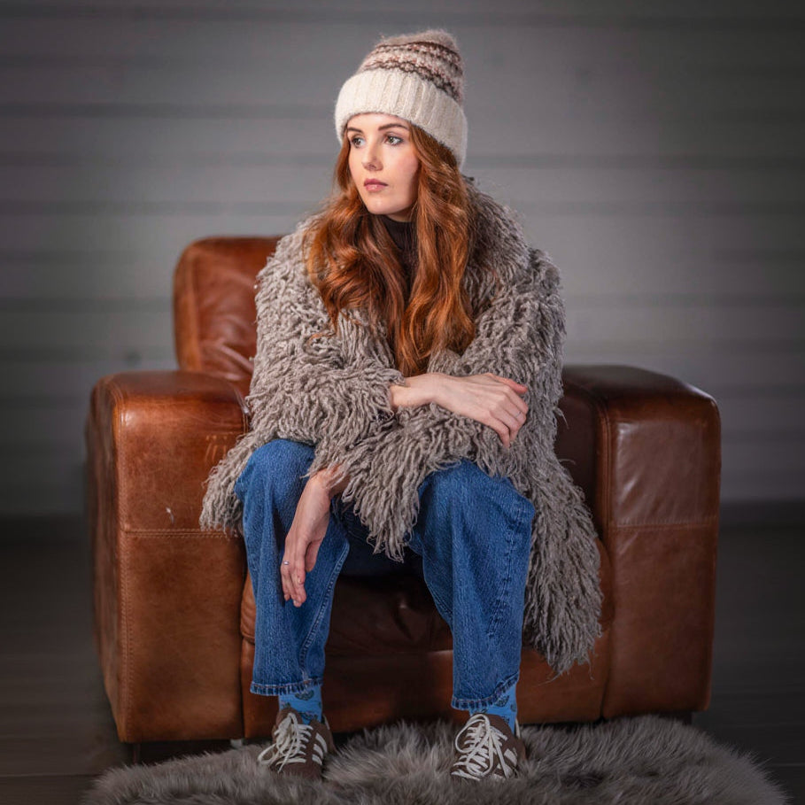 Woman sitting on a brown leather couch wearing a fluffy gray sweater, blue jeans, and a patterned hat.