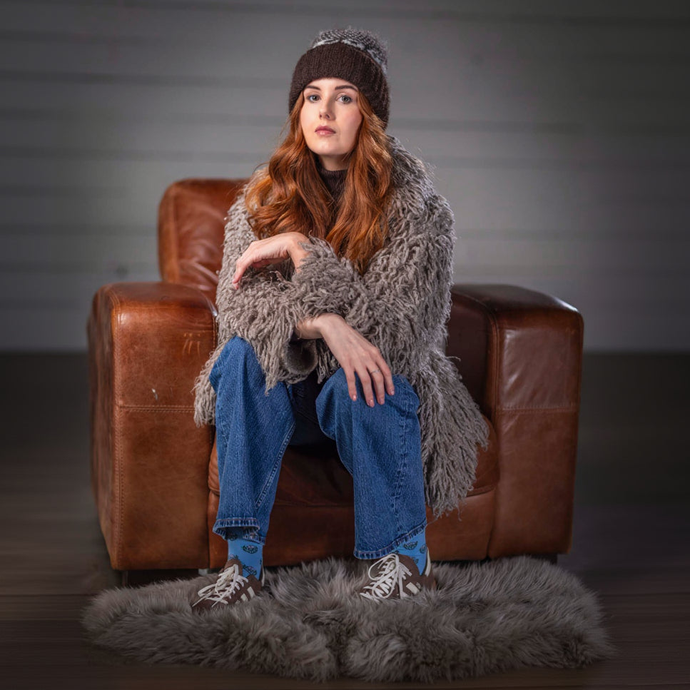 Woman sitting on a brown leather chair wearing a gray shaggy sweater, blue jeans, and a brown knit hat.