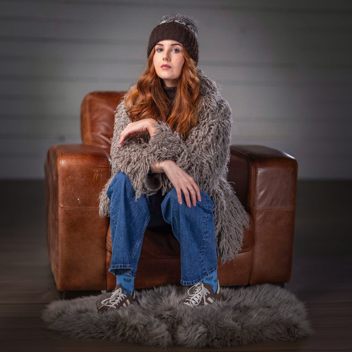 Woman sitting on a brown leather chair wearing a gray shaggy sweater, blue jeans, and a brown knit hat.