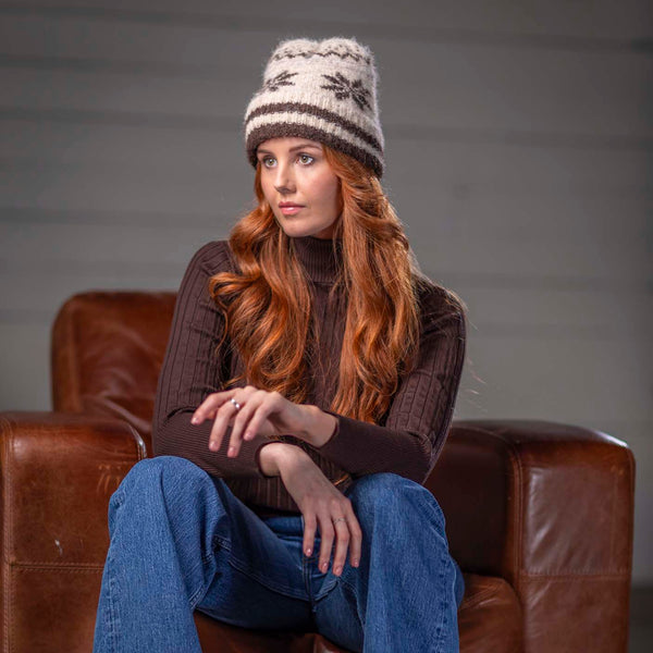 A woman with long red hair sits in a brown leather armchair wearing a cream and brown knit beanie with a snowflake pattern, a dark brown ribbed turtleneck sweater, and blue jeans.