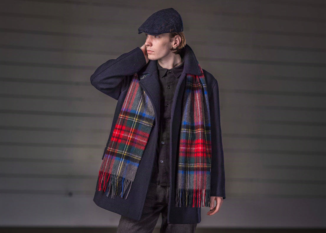 A model wearing a dark navy coat, a red and green tartan plaid scarf, flat cap, and dark trousers, standing against a grey wall.