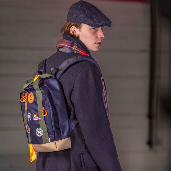Person wearing a dark coat and hat with a backpack featuring patches, standing against a plain background.