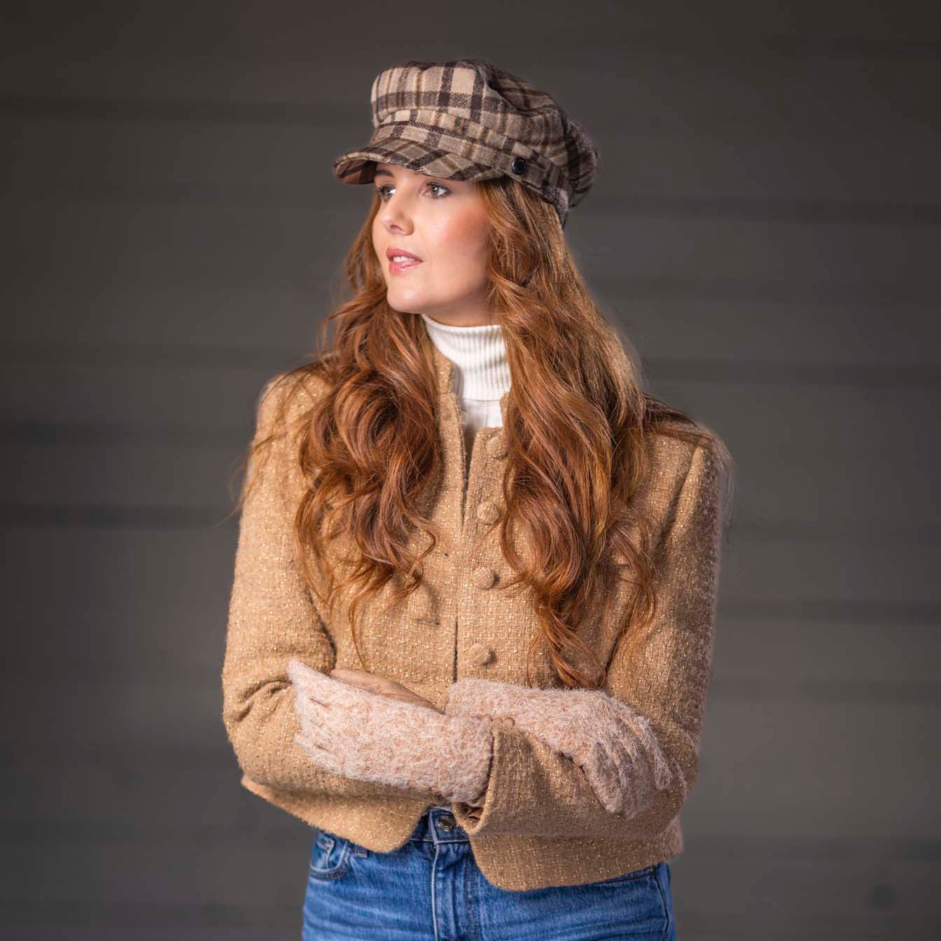 A woman wearing a beige cropped knit cardigan, blue jeans, a white turtleneck, and a brown plaid newsboy cap, standing against a dark gray background.