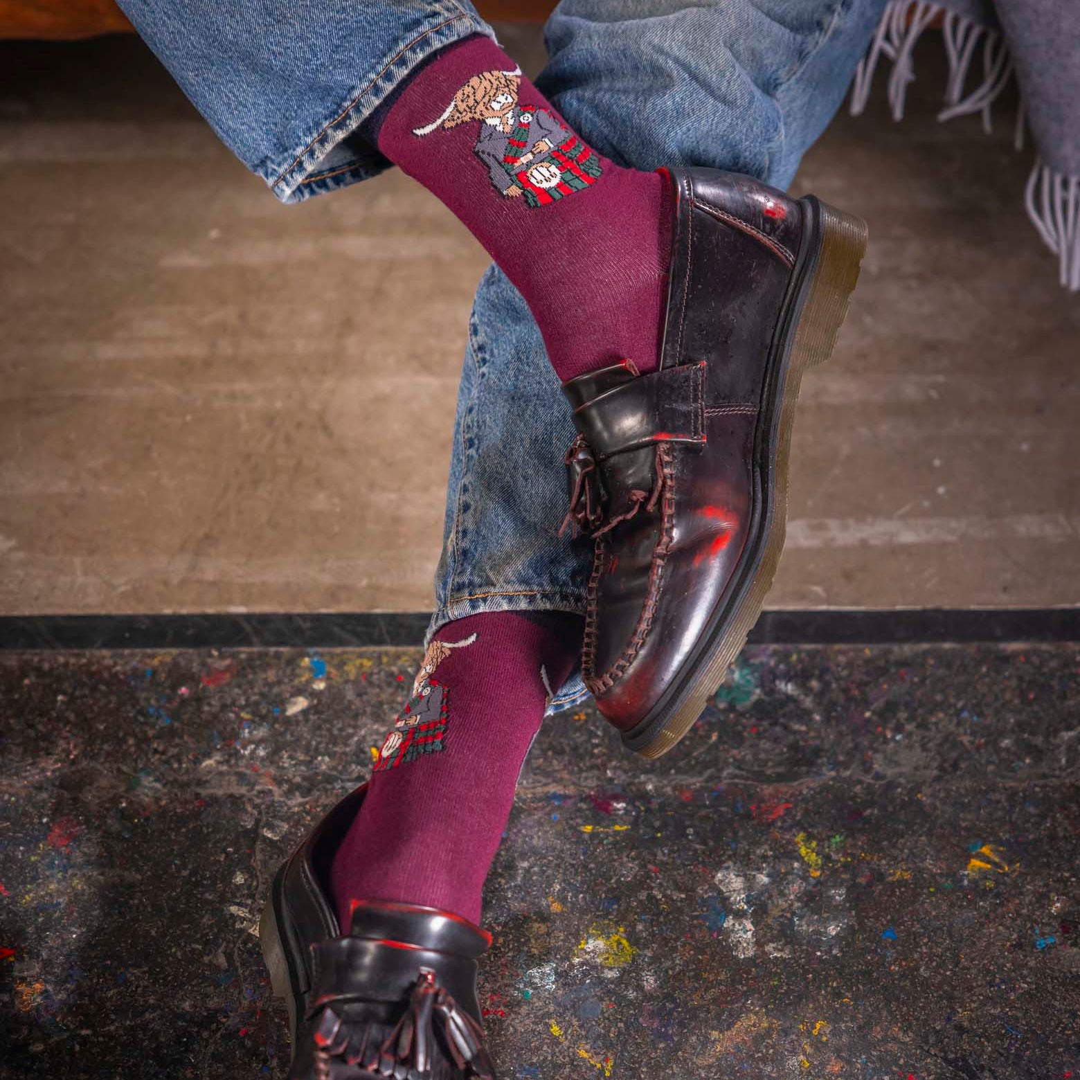Person wearing maroon socks with embroidery, blue jeans, and black shoes on a colorful floor.