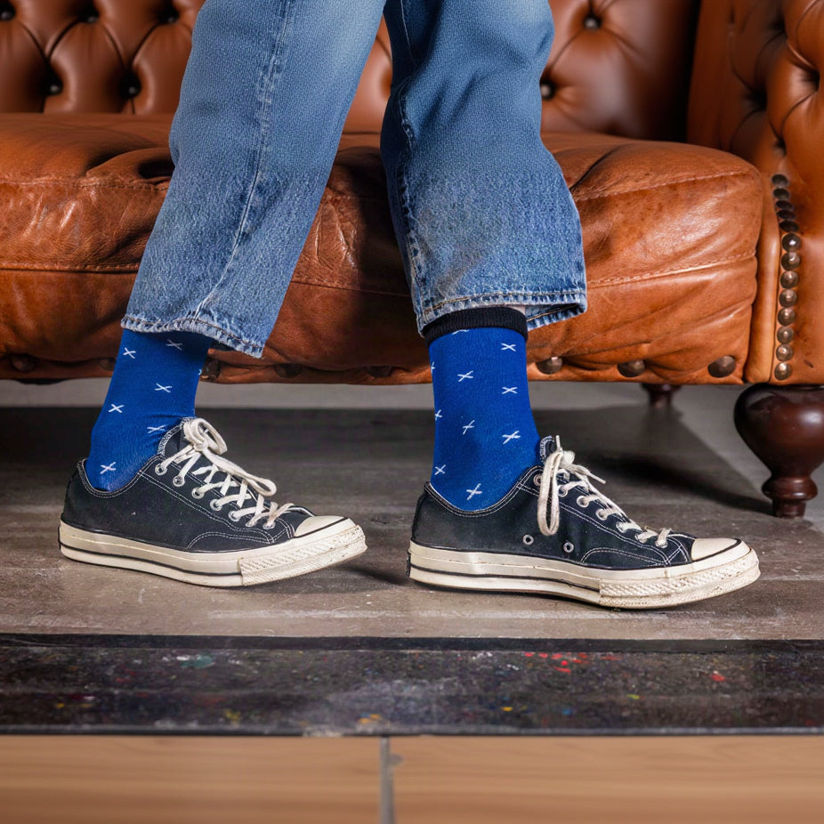 Person wearing blue socks with Scotland flag pattern and black sneakers on a wooden floor.