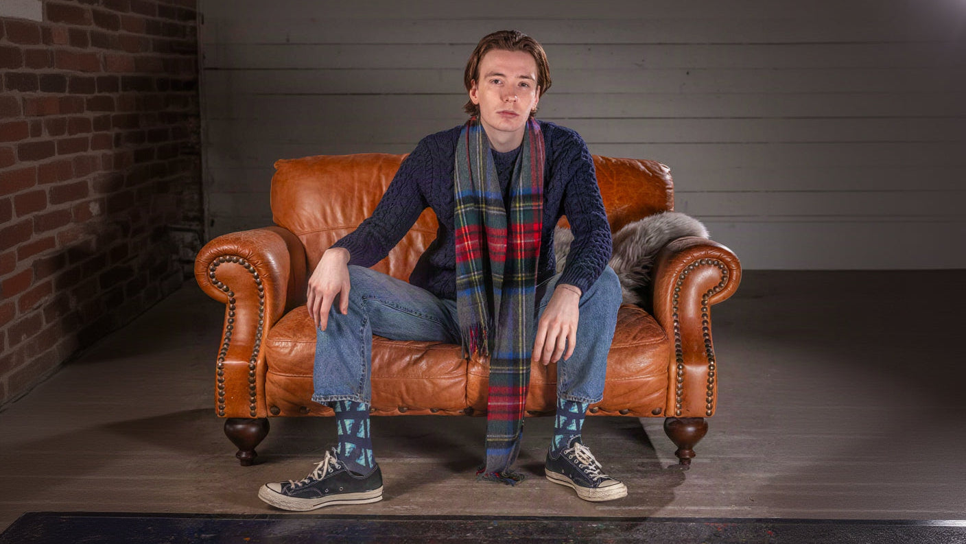 A young man sits on a brown leather armchair in a studio setting, wearing a navy sweater, a red and green plaid scarf, blue jeans, and Converse sneakers.